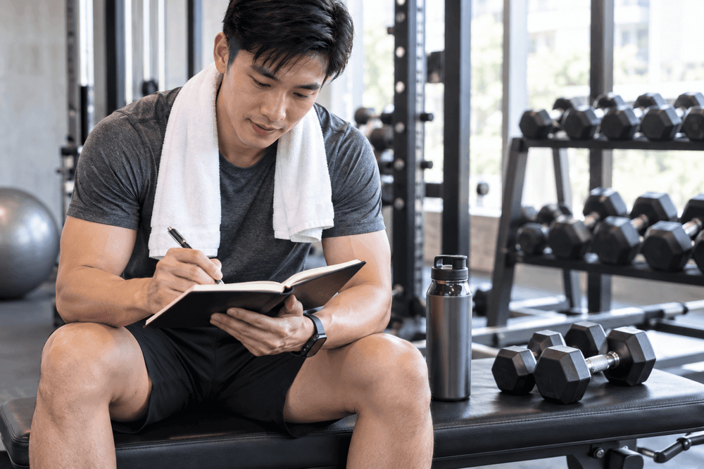 Young man sitting in a gym, writing notes in a small book with a pen.